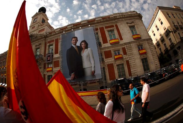 People holding Spanish flags walk past a picture showing Spain's new King Felipe VI and his wife Queen Letizia hanging on a buiding's facade in Madrid