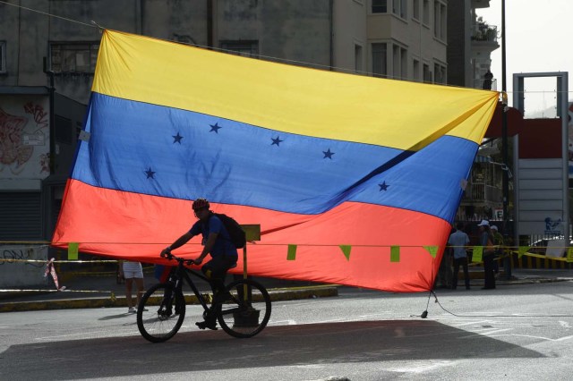 A man rides a bicycle in front of a Venezuelan national flag in Caracas on July 10, 2017. Venezuela hit its 100th day of anti-government protests Sunday, amid uncertainty over whether the release from prison a day earlier of prominent political prisoner Leopoldo Lopez might open the way to negotiations to defuse the profound crisis gripping the country. / AFP PHOTO / Federico Parra